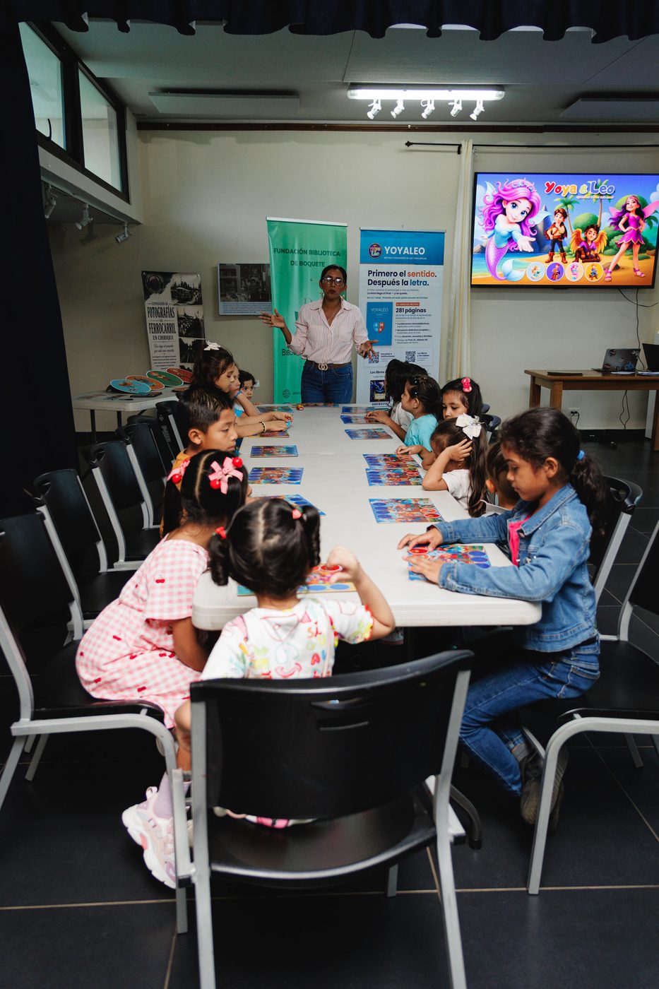 La sala del taller con los niños en la mesa larga y la pantalla de Yoya y Leo al fondo.