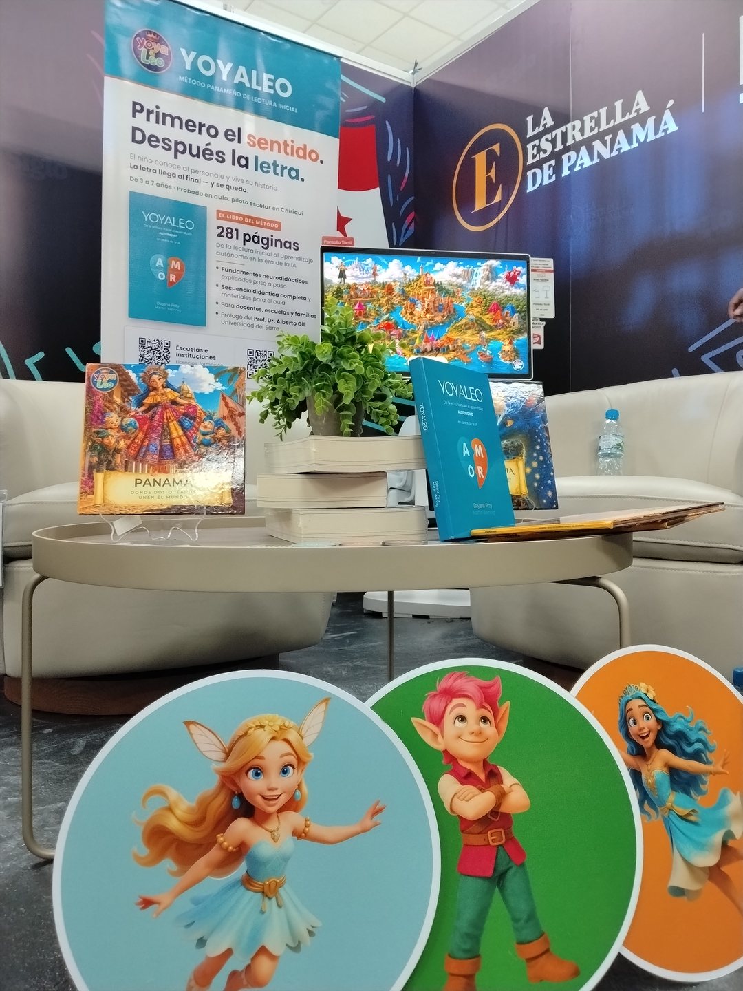 El estand de Yoya & Leo en la Feria Internacional del Libro de Panamá 2026: los dos libros impresos, el mapa de Amoravia en pantalla y las fichas de los personajes